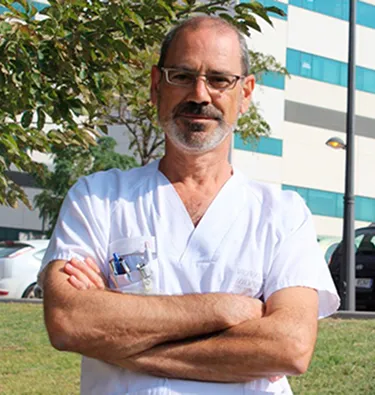 Hombre con gafas y barba, canoso con brazos cruzados y camiseta de médico blanca con boligrafos en el bolsillo en el exterior de un hospital