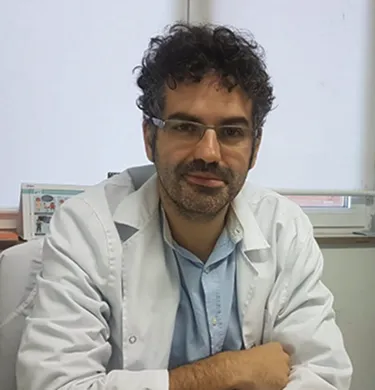 Hombre con gafas y barba de pelo oscuro con bata blanca sobre camisa azul, sentado. 