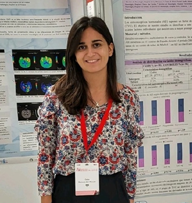 Mujer morena sonriente de pelo oscuro con camisa estampada de flores con una tarjeta identificativa sobre un fondo de presentaciones de proyectos. 