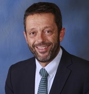 Hombre con gafas y barba blanca de pelo oscuro con bata blanca sobre camisa azul, sentado. 