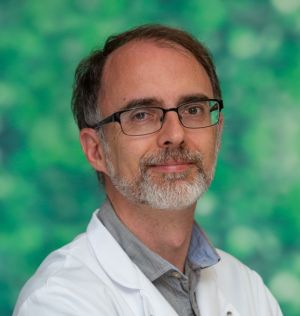 Hombre con gafas y barba blanca de pelo oscuro con bata blanca sobre camisa azul, sentado.